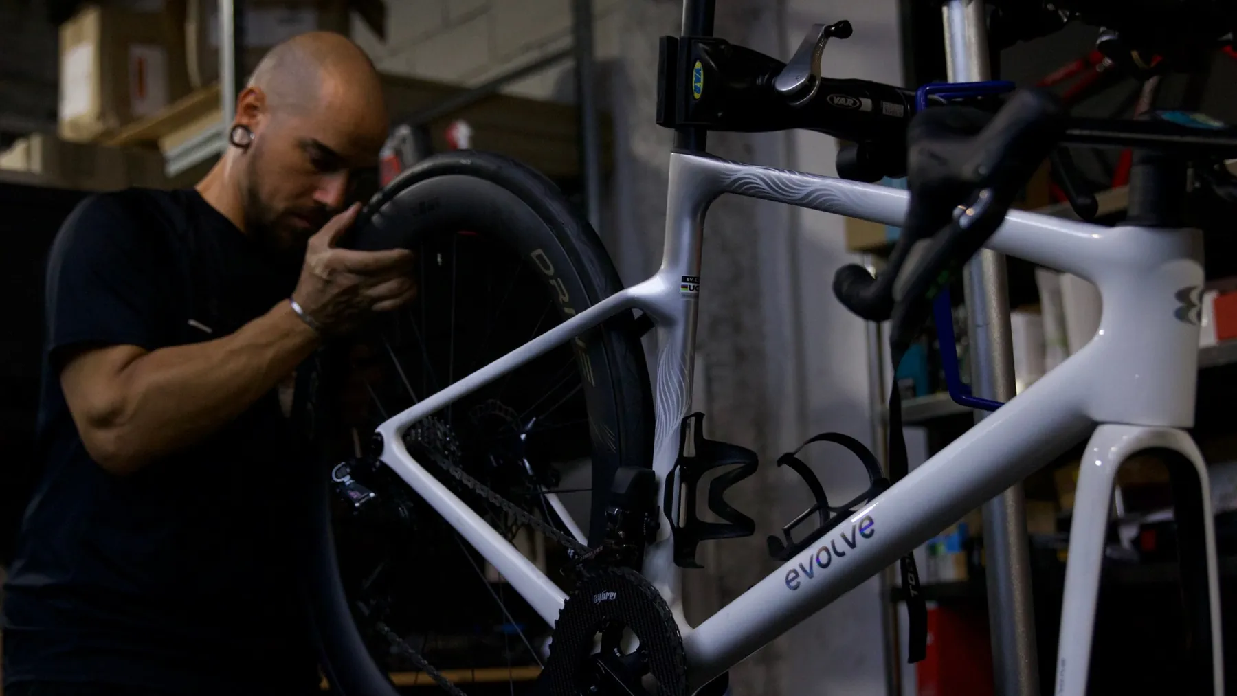 In the workshop, a mechanic checks the rear wheel of a white carbon road bike before fitting it to the frame on the stand.