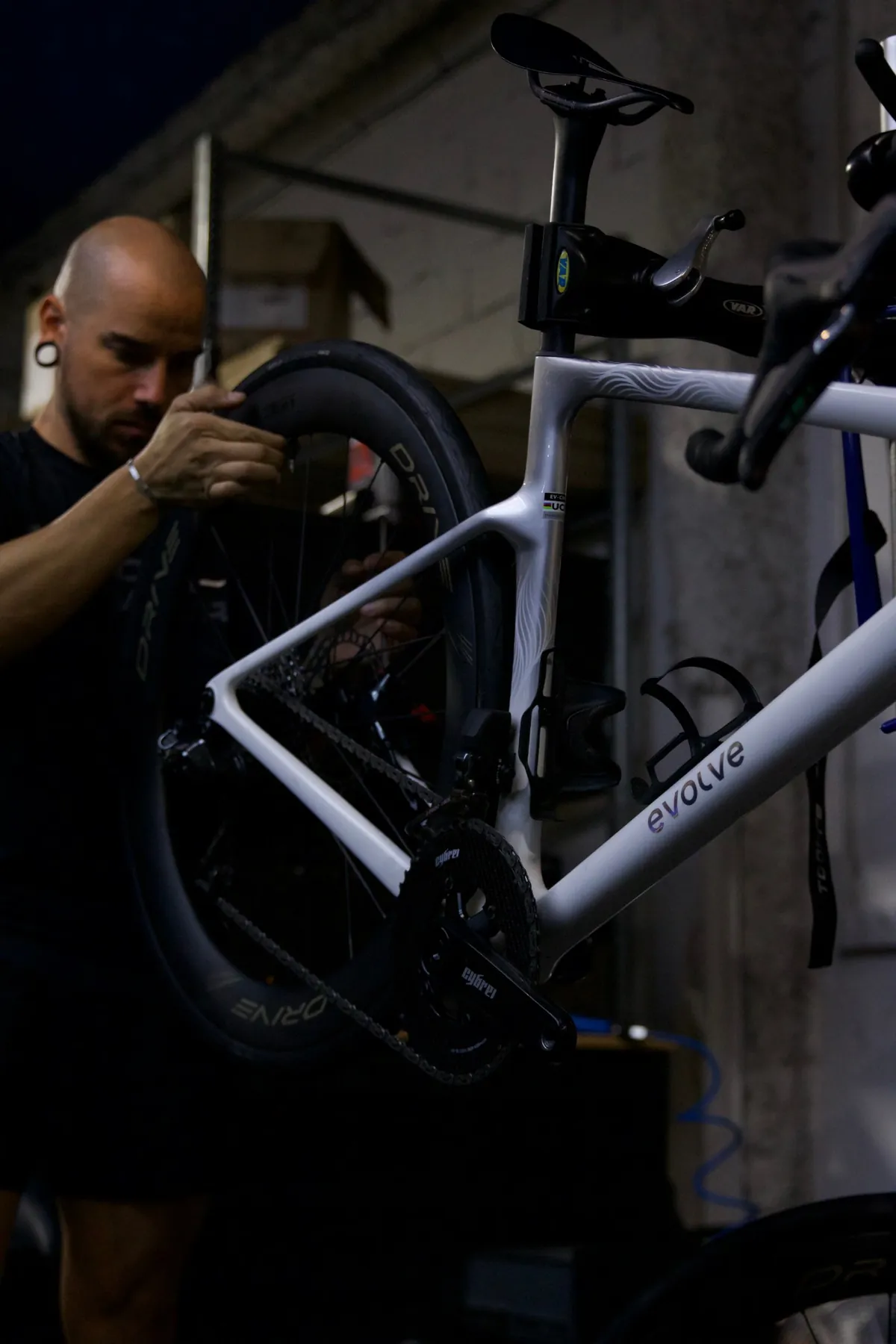 A mechanic fits the rear wheel of a white carbon road bike held in the workshop stand.