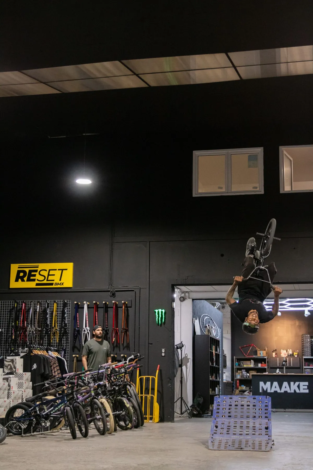 A BMX rider flips over a ramp on the shop floor, in front of the Reset BMX wall and the MAAKE counter.